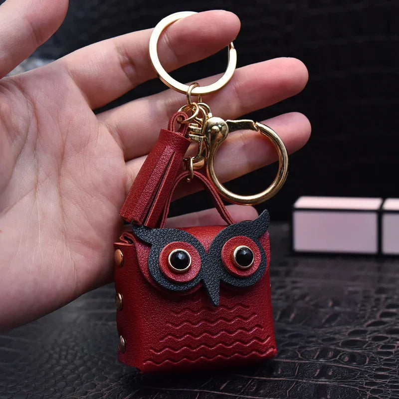 Hand holding red leather owl keychain with black eyes and gold keyring on textured black surface