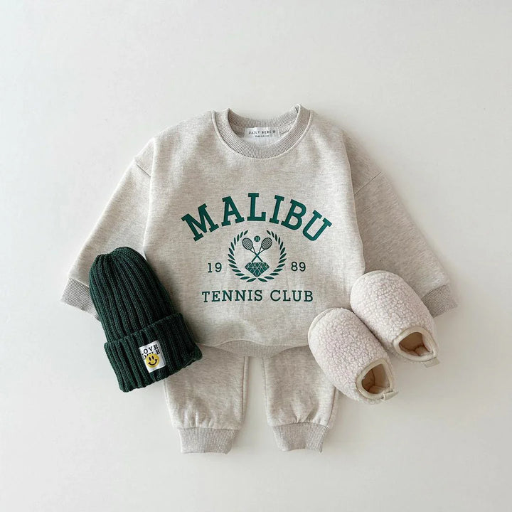Toddler beige sweatshirt and pants set with Malibu tennis club print, green knit hat, and cream slippers