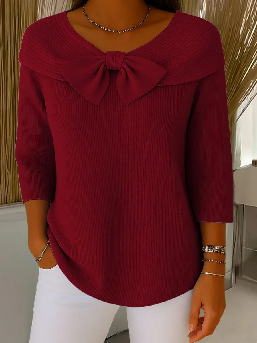 Woman wearing burgundy ribbed sweater with large bow detail and white pants, accessorized with silver jewelry