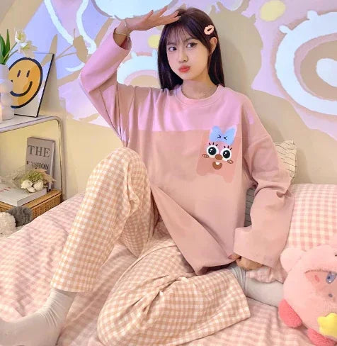 Young woman in pink cartoon sweatshirt and checkered pajama pants sitting on bed with pastel decor
