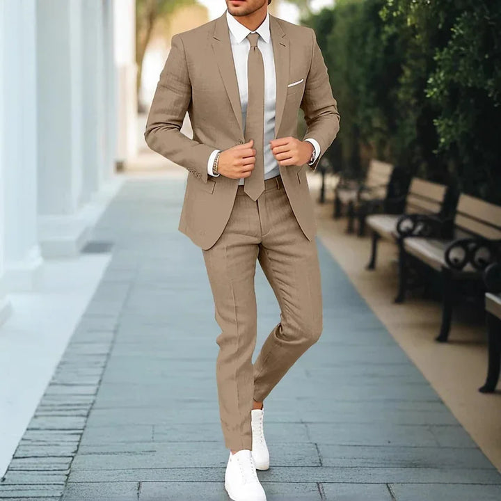 Man in beige slim-fit suit with white shirt and tie walking on outdoor pathway with benches