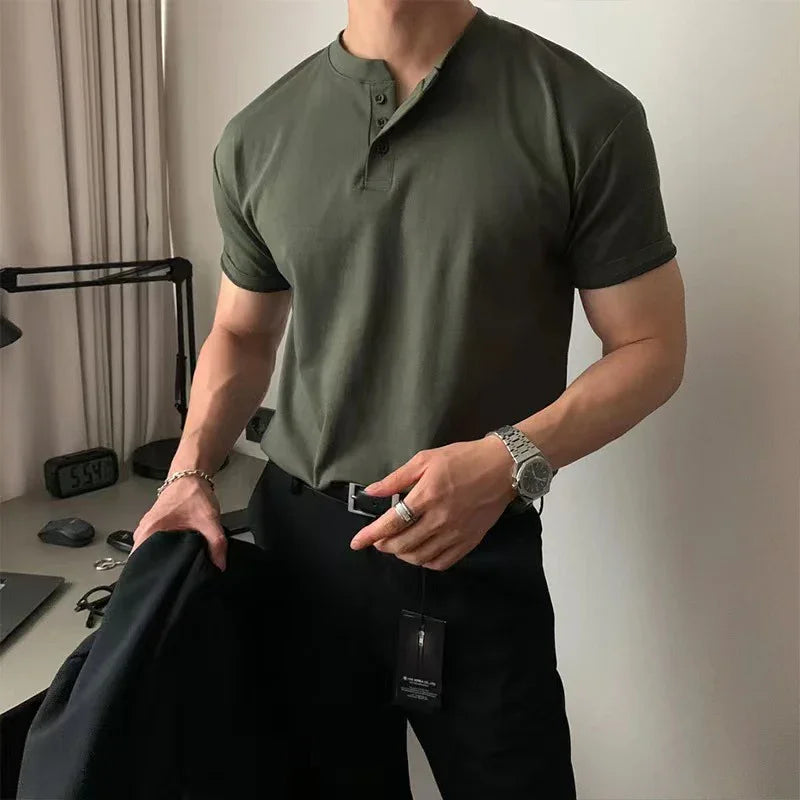 Man wearing fitted olive green henley t-shirt and black pants with watch holding black jacket indoors