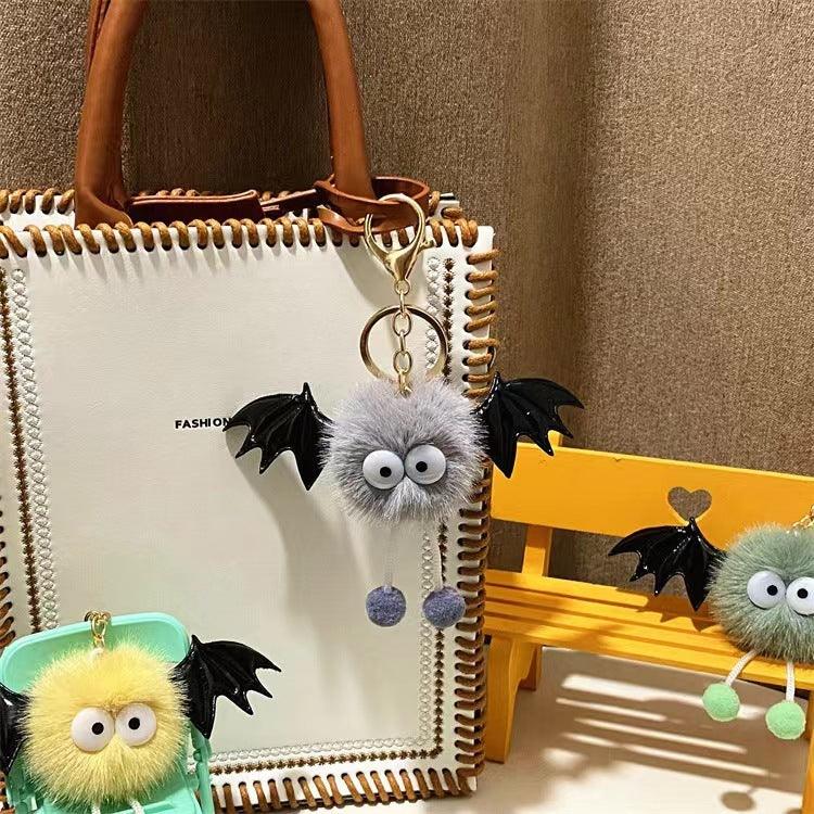 Furry bat keychains with large eyes and black wings attached to a white handbag and yellow miniature chair