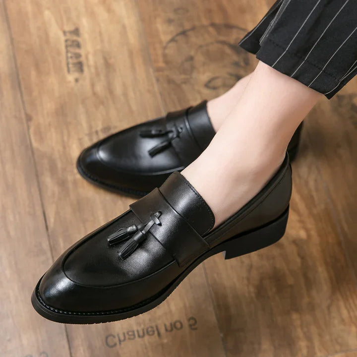 Black leather tassel loafers worn with pinstripe pants on wooden floor