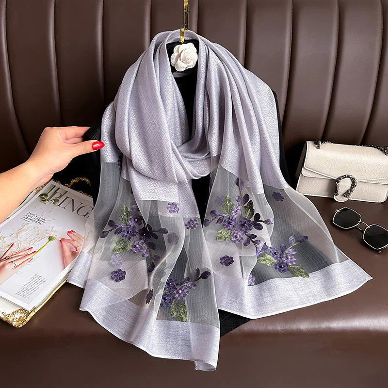 Elegant silver-gray scarf with sheer purple floral embroidery displayed on black mannequin against brown leather background