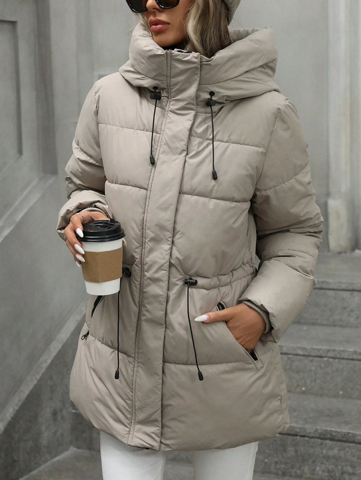 Woman wearing beige padded winter jacket with high collar and drawstrings holding coffee cup