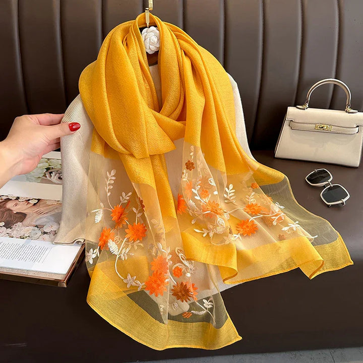 Yellow scarf with floral embroidery draped on beige sweater, next to beige handbag and black sunglasses