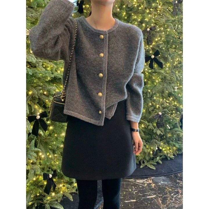 Woman wearing a gray buttoned cardigan and black skirt standing in front of a decorated Christmas tree with lights and black bows