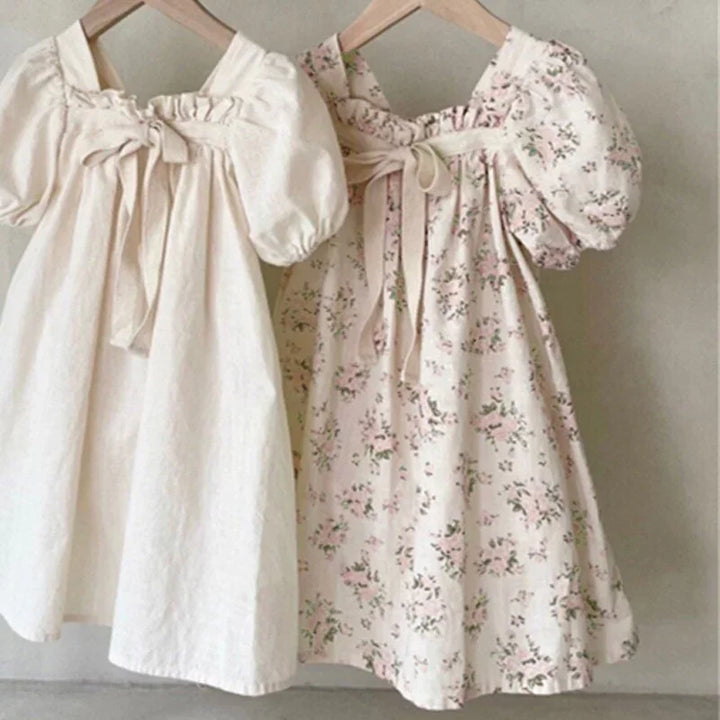 Two puff-sleeve girls' dresses, one plain cream and one cream with pink floral print, hanging on wooden hangers