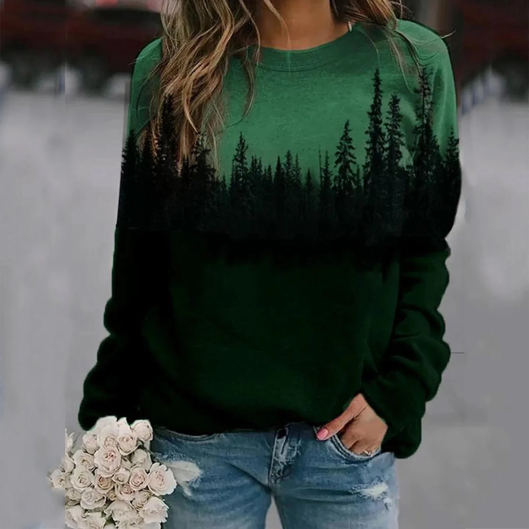 Woman wearing green sweatshirt with black forest tree line print, holding white flowers, paired with distressed jeans