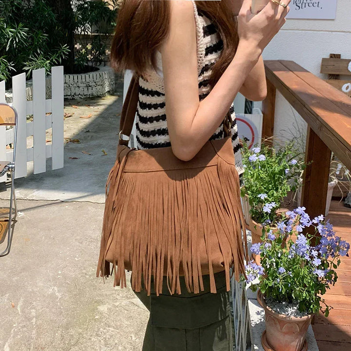 Woman wearing a black and white striped top holding a brown fringe shoulder bag outdoors near potted purple flowers