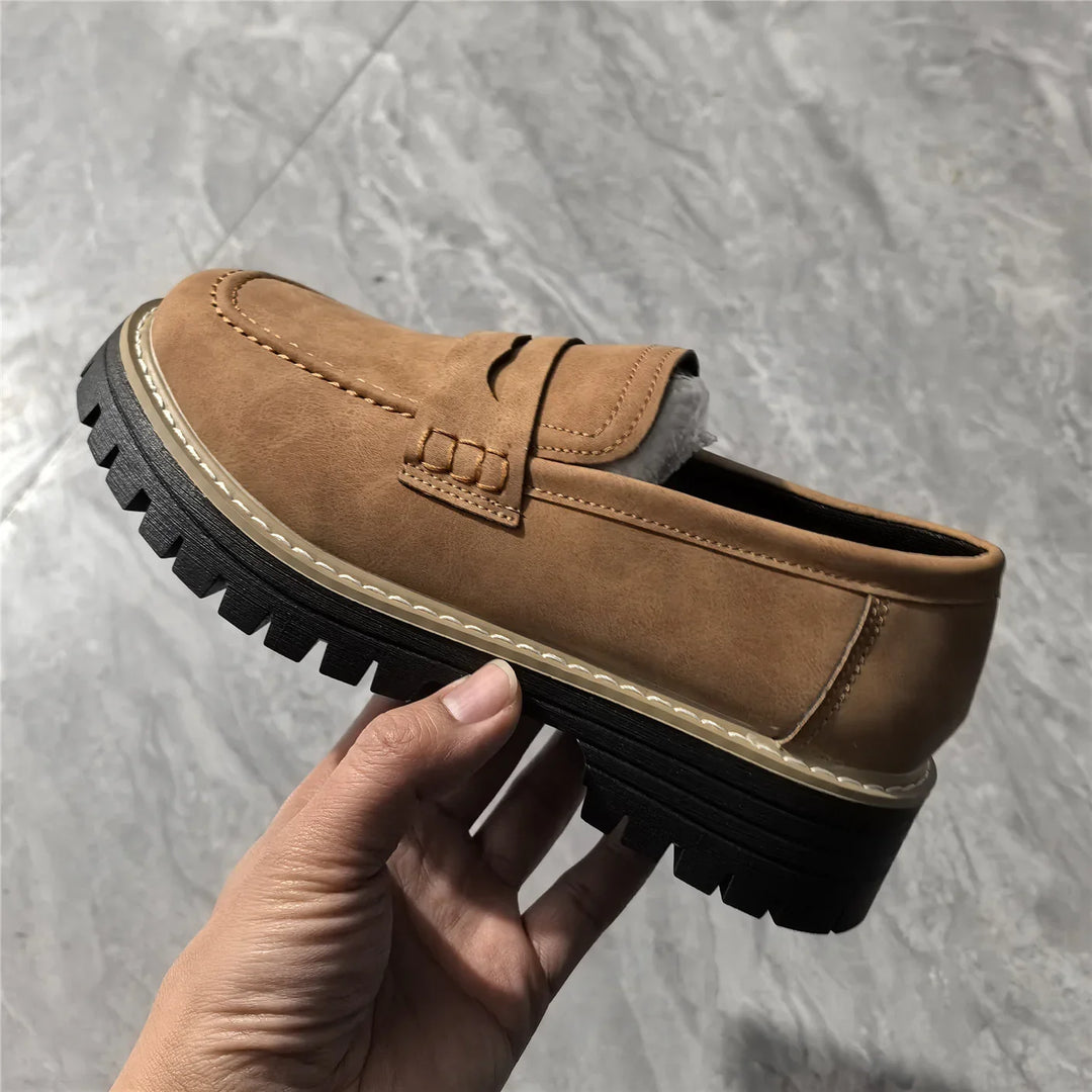 Hand holding a tan brown leather loafer with black chunky sole and white stitching on marble floor