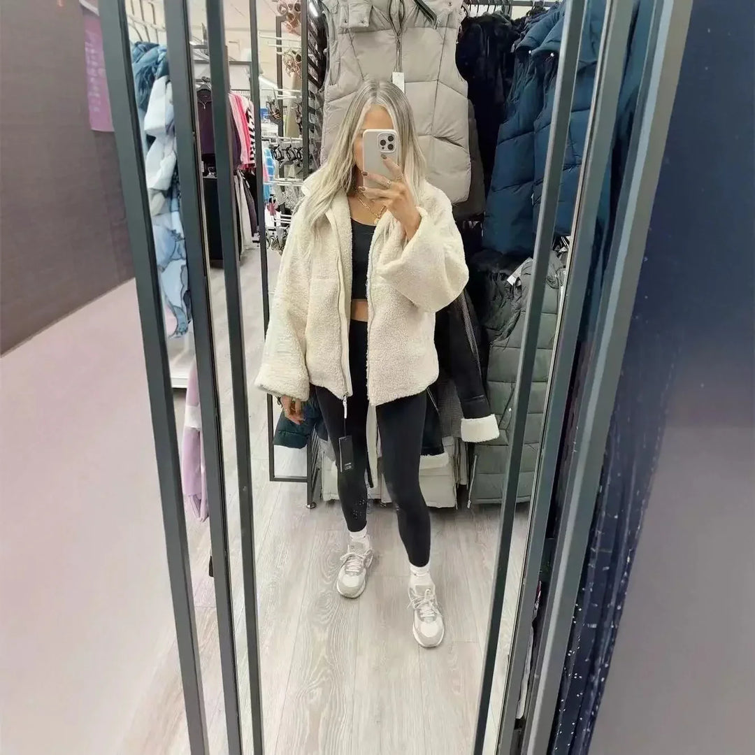 Woman in white fuzzy jacket, black leggings, and sneakers taking mirror selfie in clothing store