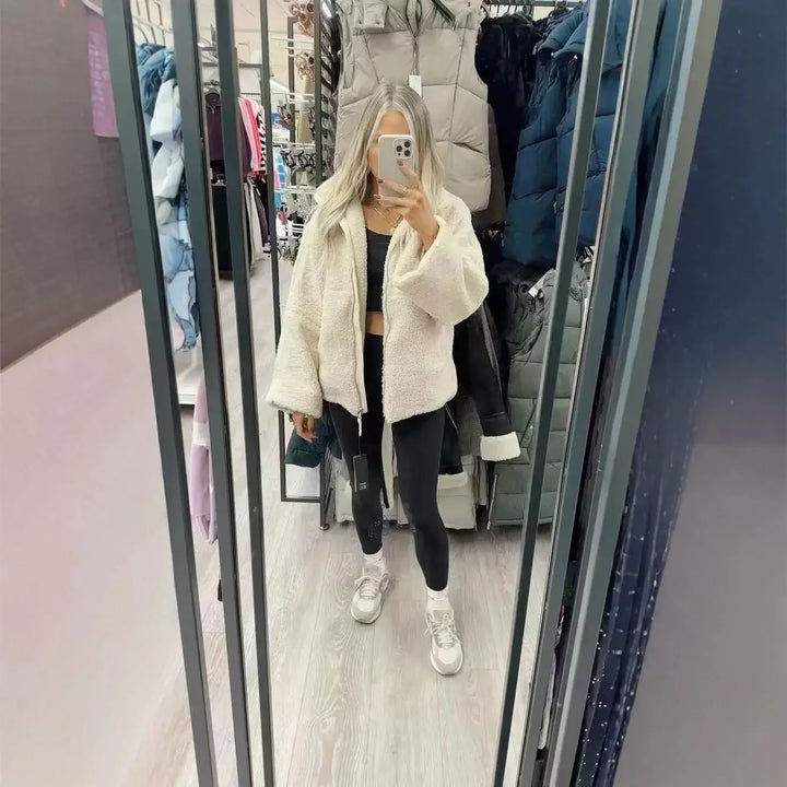 Woman in white fuzzy jacket, black leggings, and sneakers taking mirror selfie in clothing store