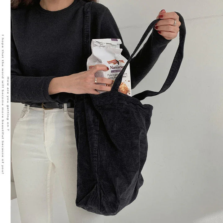 Woman wearing black sweater and white pants holding black corduroy tote bag with packaged cookies inside