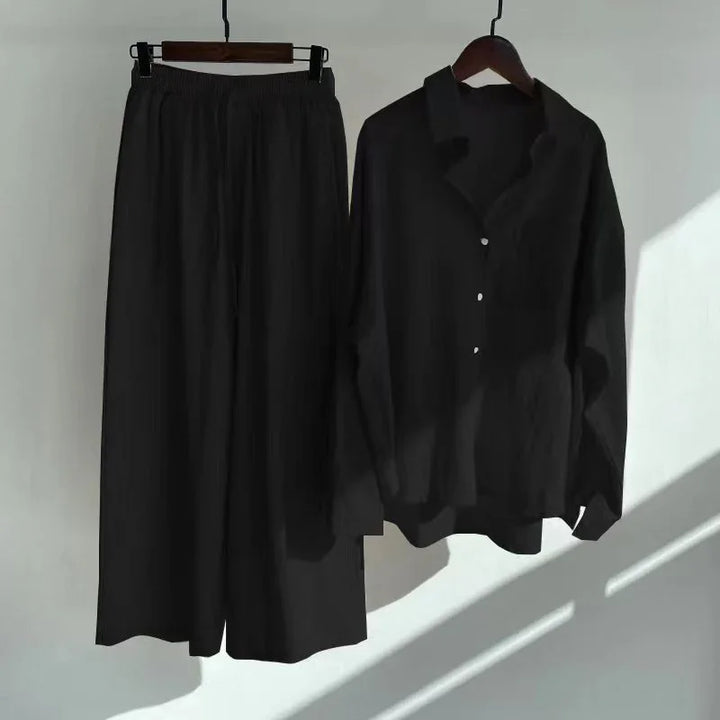 Black loose pants with elastic waist and matching black button-up shirt on hangers against white wall