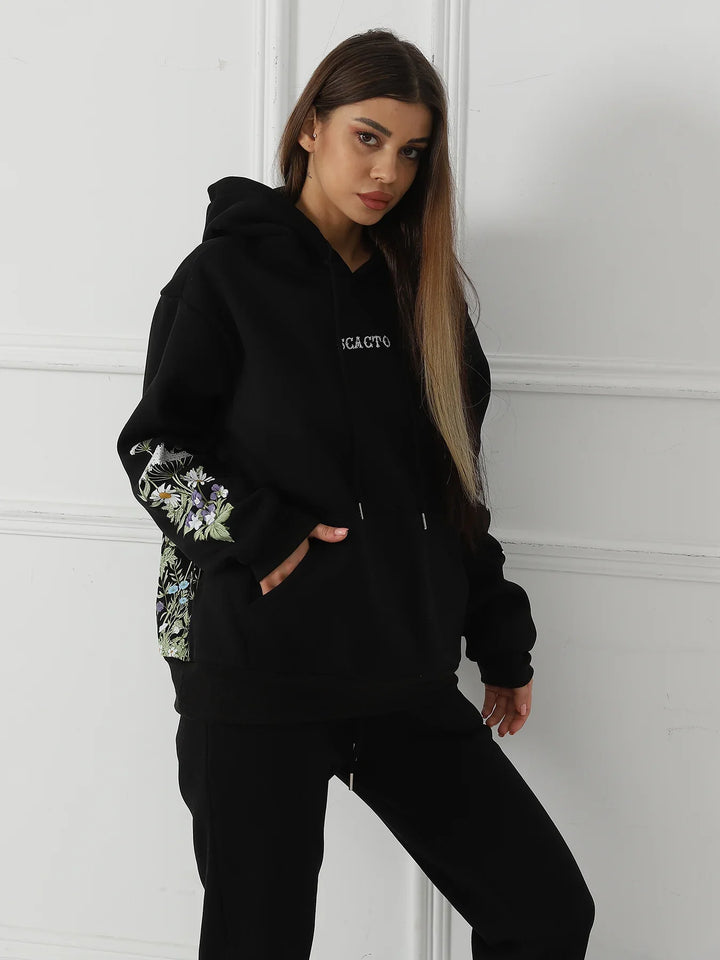 Young woman wearing black hoodie with floral embroidery on sleeves and black pants against white wall