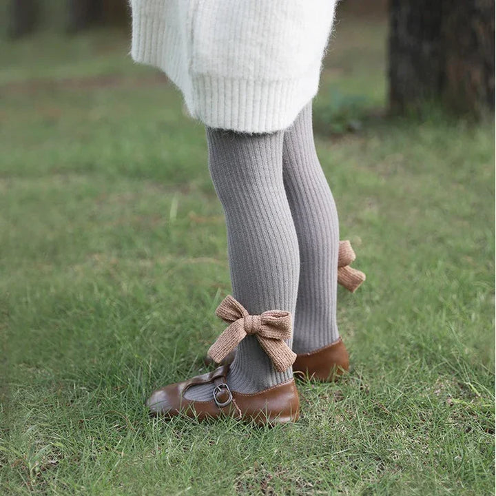 Child wearing gray ribbed tights with brown knit bows and brown leather Mary Jane shoes on grass