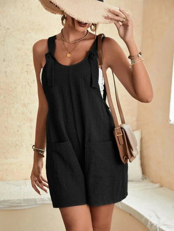 Woman in black sleeveless romper with front pockets, beige sun hat, gold jewelry, and brown shoulder bag