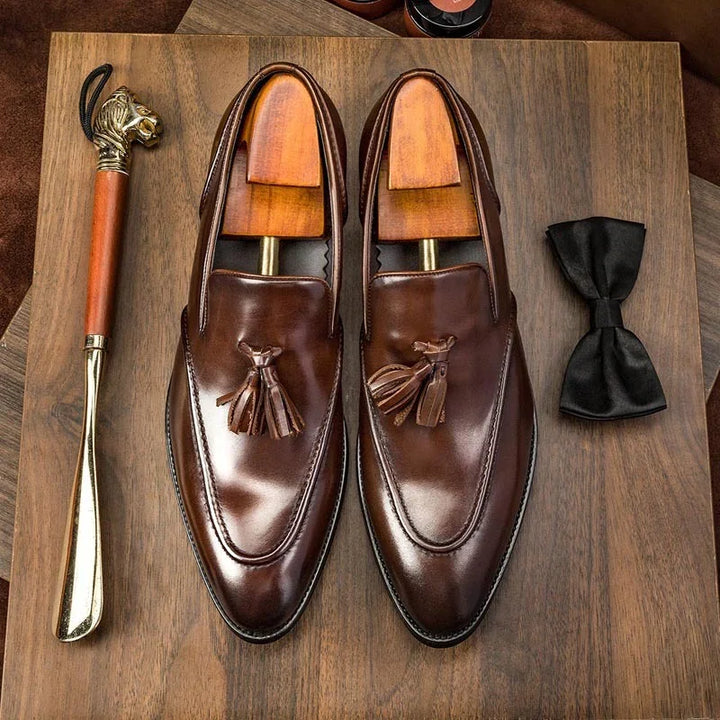Brown leather tassel loafers with wooden shoe trees, black bow tie, and tiger head shoehorn on wood surface