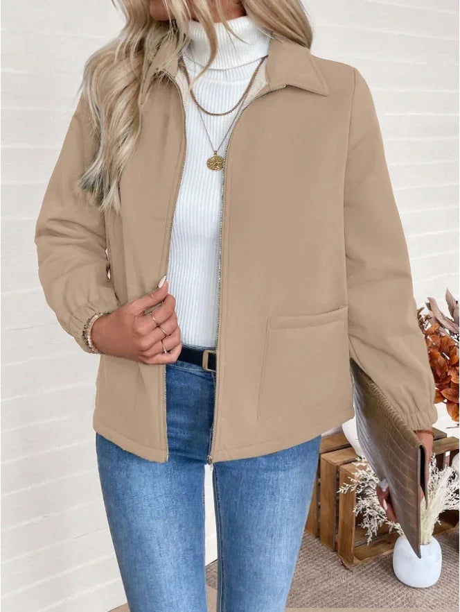 Woman wearing beige zip-up jacket over white turtleneck and blue jeans, holding brown clutch