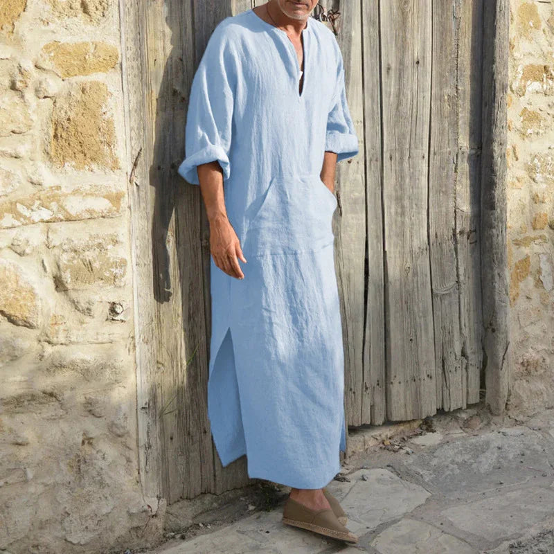 Man wearing light blue linen kaftan with rolled sleeves standing against rustic wooden door and stone wall