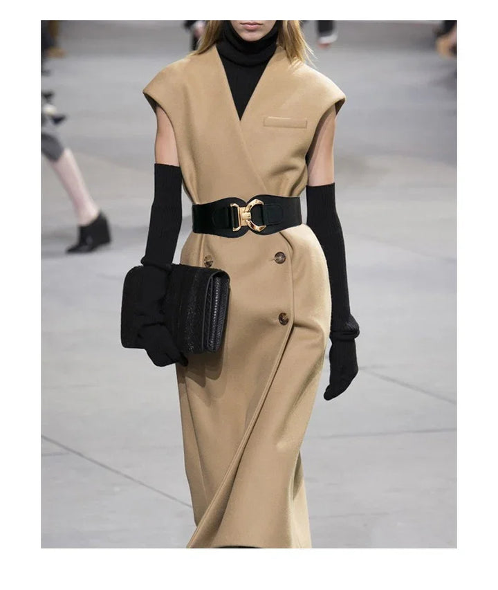 Model walking runway in beige sleeveless coat dress with black belt, turtleneck, long gloves, and clutch