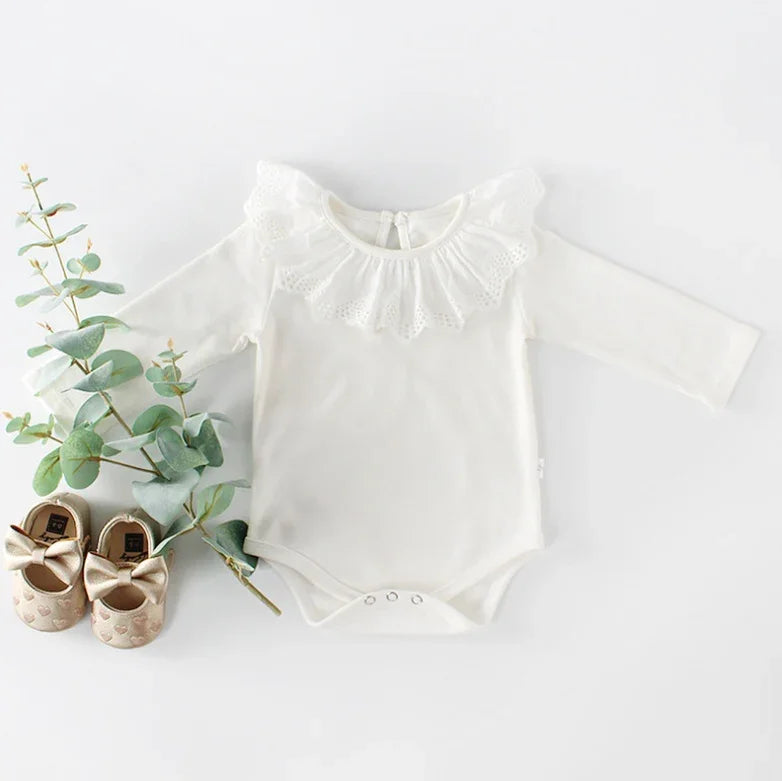 White baby bodysuit with lace collar and long sleeves, paired with gold bow baby shoes and eucalyptus branch