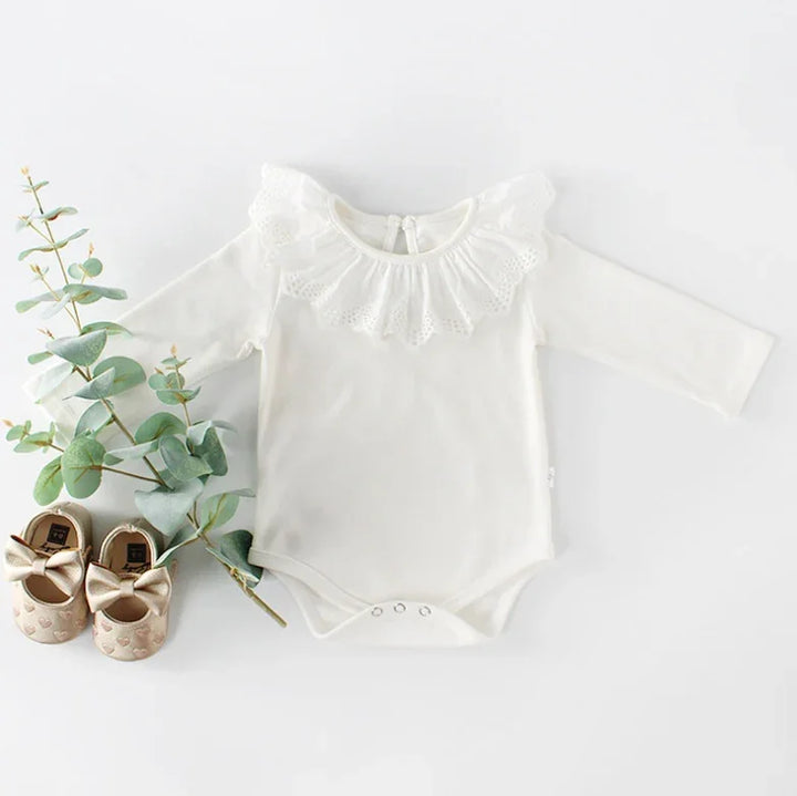 White baby bodysuit with lace collar and long sleeves, paired with gold bow baby shoes and eucalyptus branch