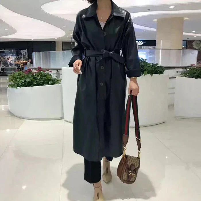 Woman wearing black belted leather trench coat holding brown designer handbag in modern mall