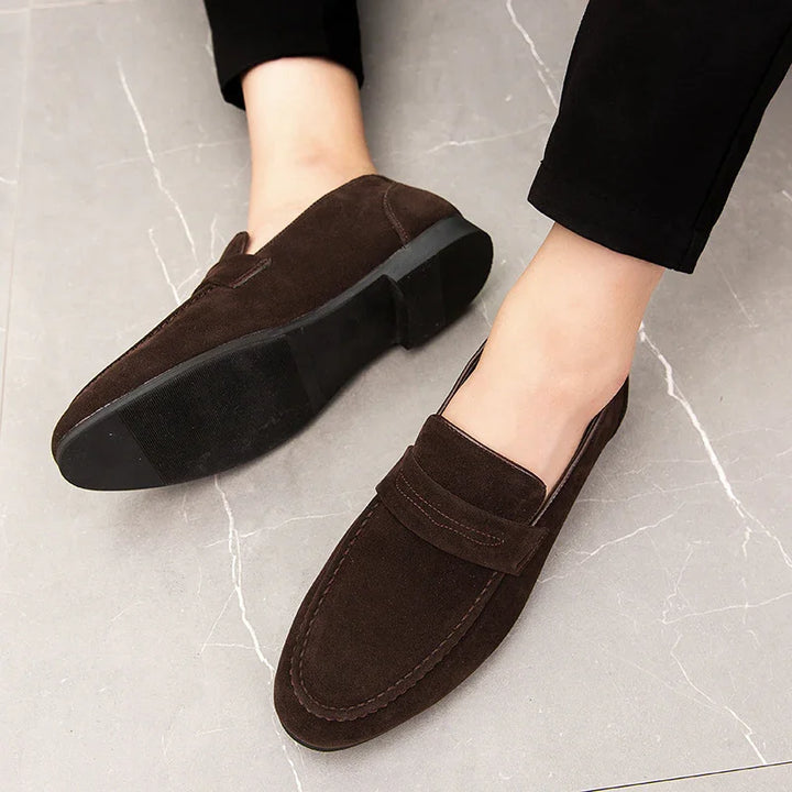 Dark brown suede men's loafers with black rubber soles worn with black pants on light gray tiled floor