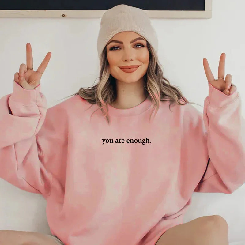 Smiling woman wearing beige beanie and pink sweatshirt with 'you are enough' text showing peace signs