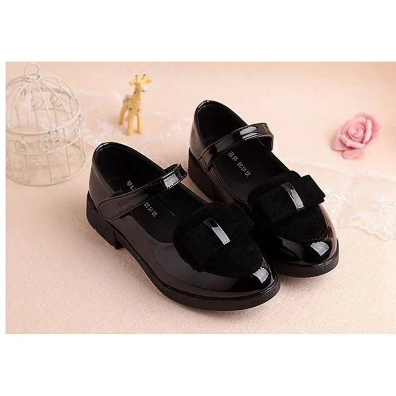 Black patent leather Mary Jane shoes with velvet bows on beige surface, decorative cage and small giraffe figure