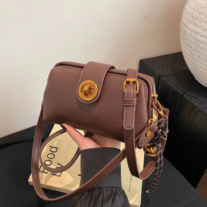 Hand holding a brown leather shoulder bag with gold buckle and a decorative charm in an indoor setting