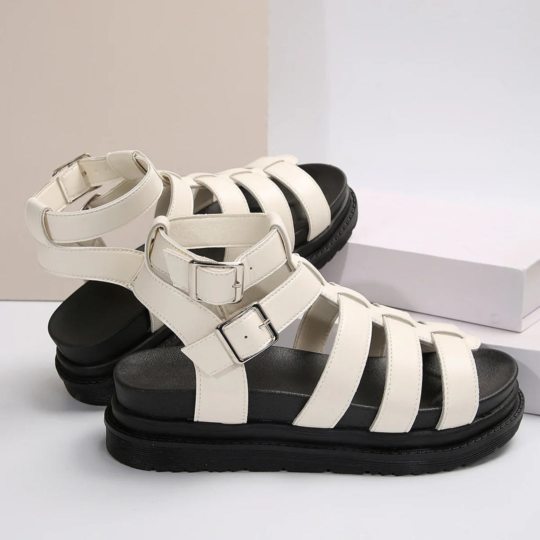 White strappy sandals with black soles and adjustable silver buckles on a white and beige background