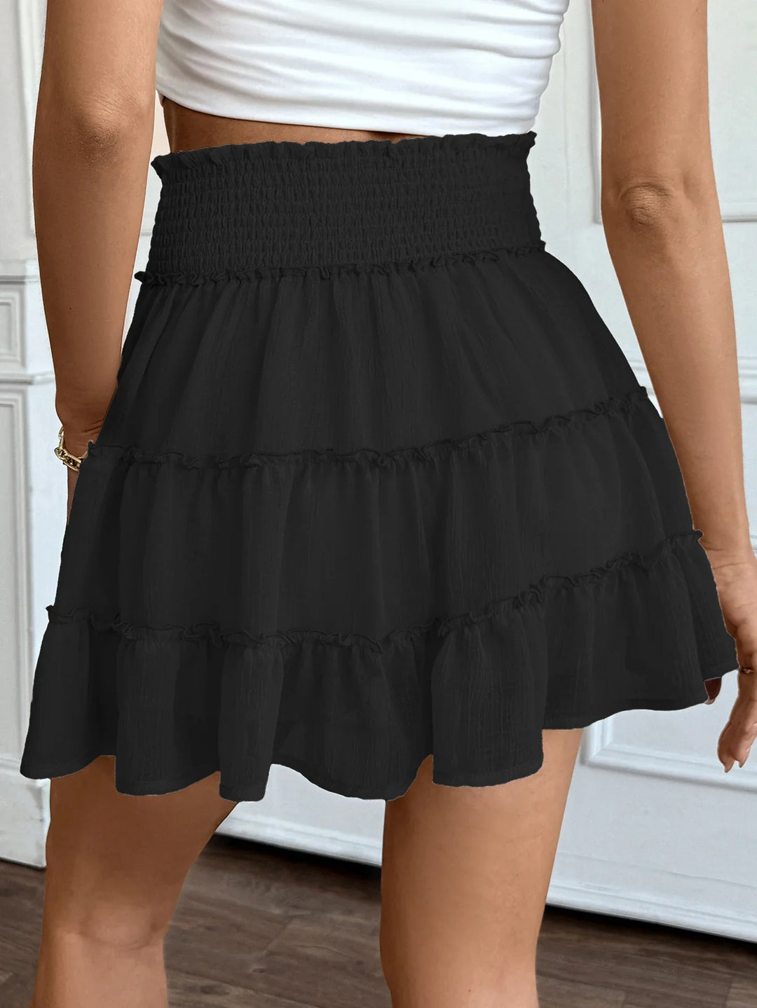 Rear view of woman wearing black ruffled mini skirt with smocked high waist and white crop top indoors