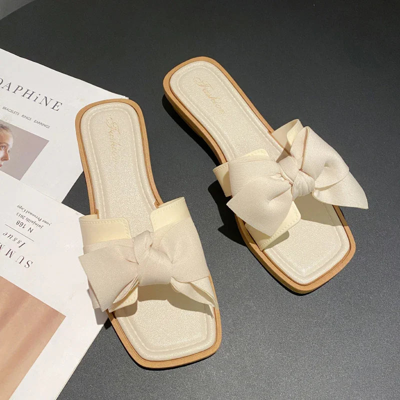Cream-colored women's flat slide sandals with large fabric bows on black surface next to an open magazine