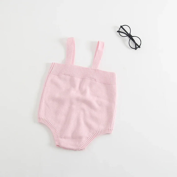 Soft pink knitted baby romper with shoulder straps on white background beside black round eyeglasses