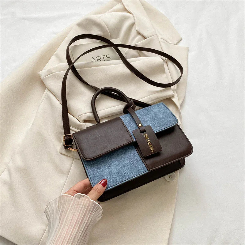 Hand holding blue and brown leather crossbody handbag over cream blazer jacket on white surface