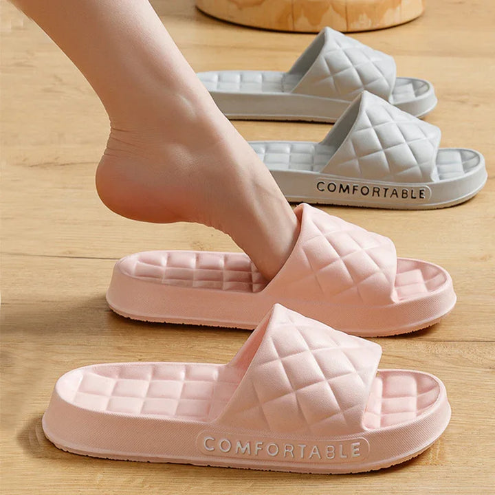 Foot slipping into pink quilted slide slippers with thick soles labeled comfortable on wooden floor, gray pair in background