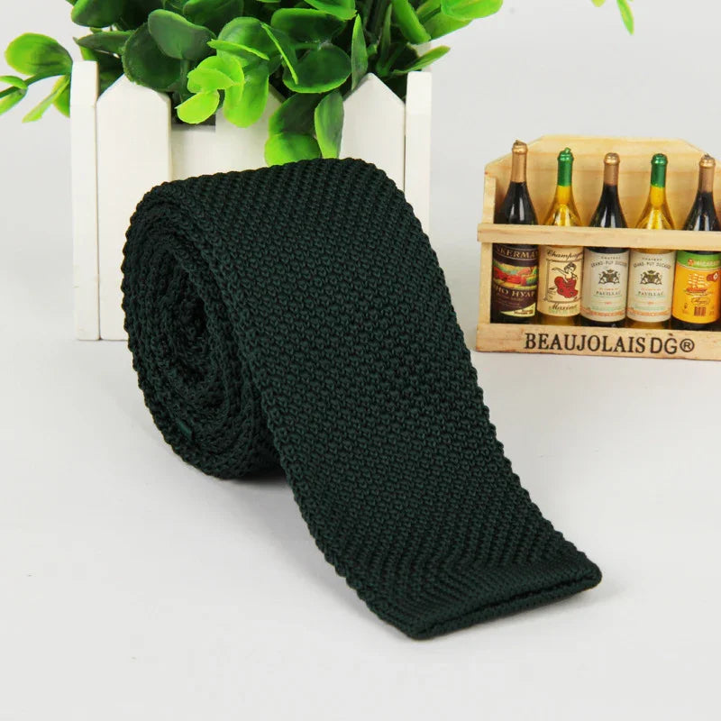 Rolled dark green knitted tie on white surface with green plant and miniature wine bottles in background