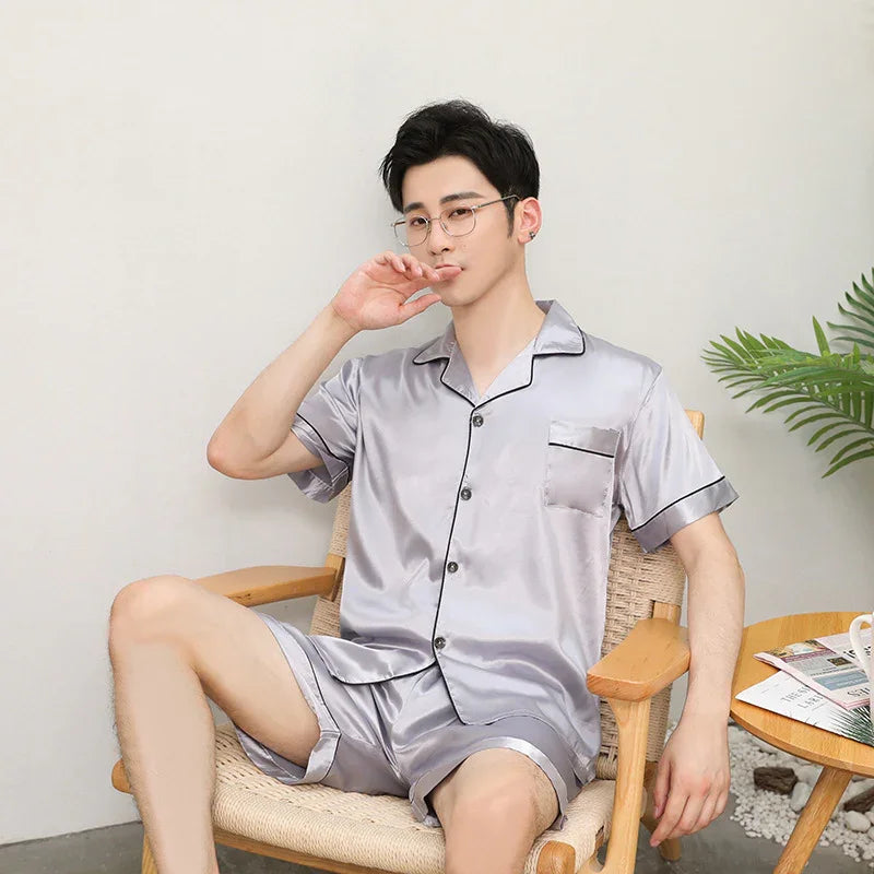 Man wearing silver satin short-sleeve pajamas sitting on woven chair beside wooden table with plant