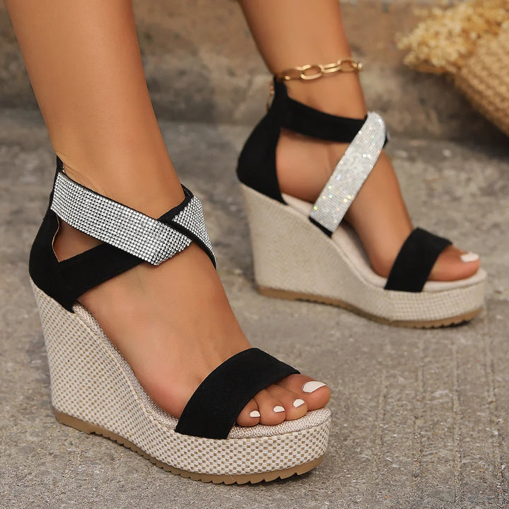 Black wedge sandals with sparkling rhinestone ankle straps on textured stone surface