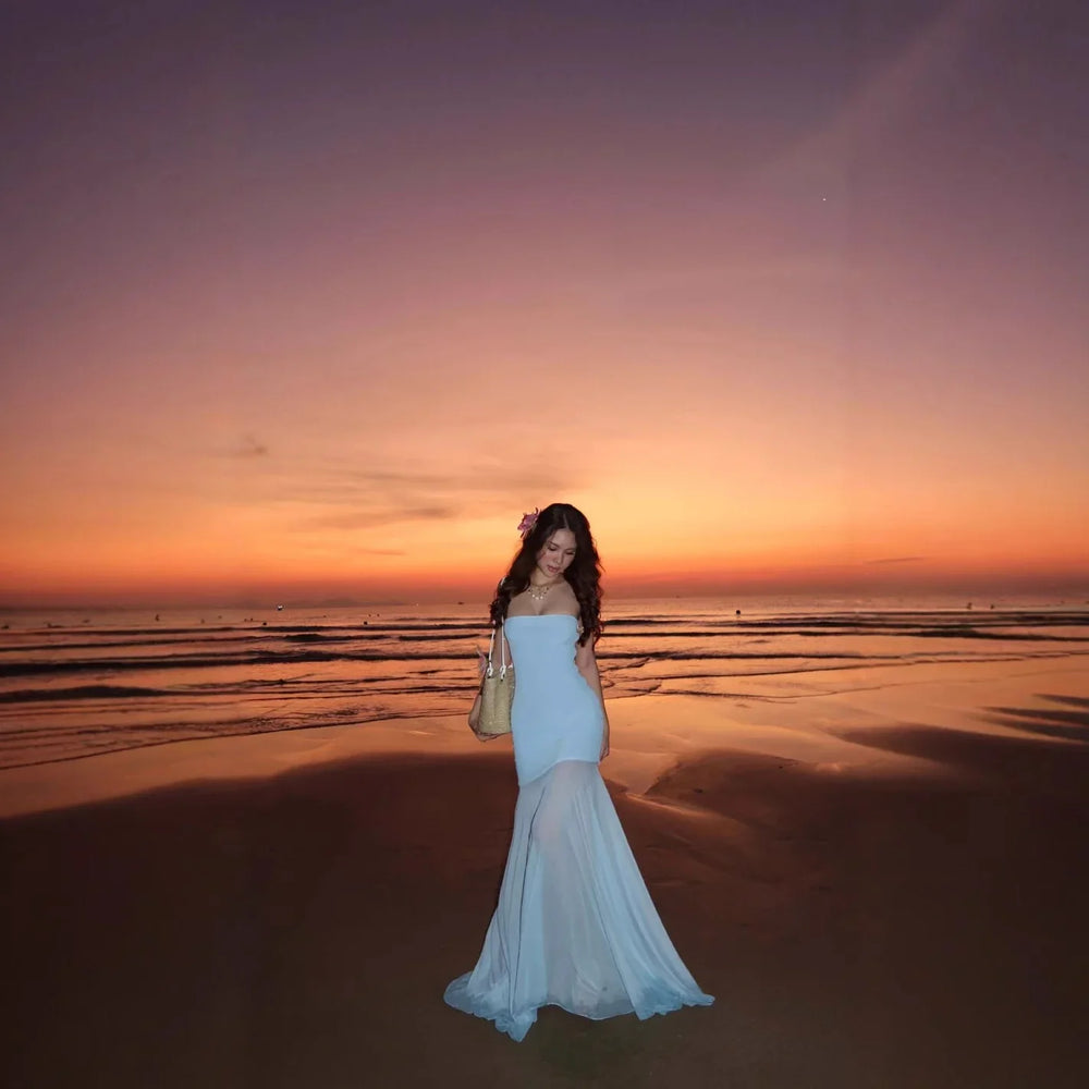 Woman in a flowing white dress standing on a beach at sunset with orange and purple sky