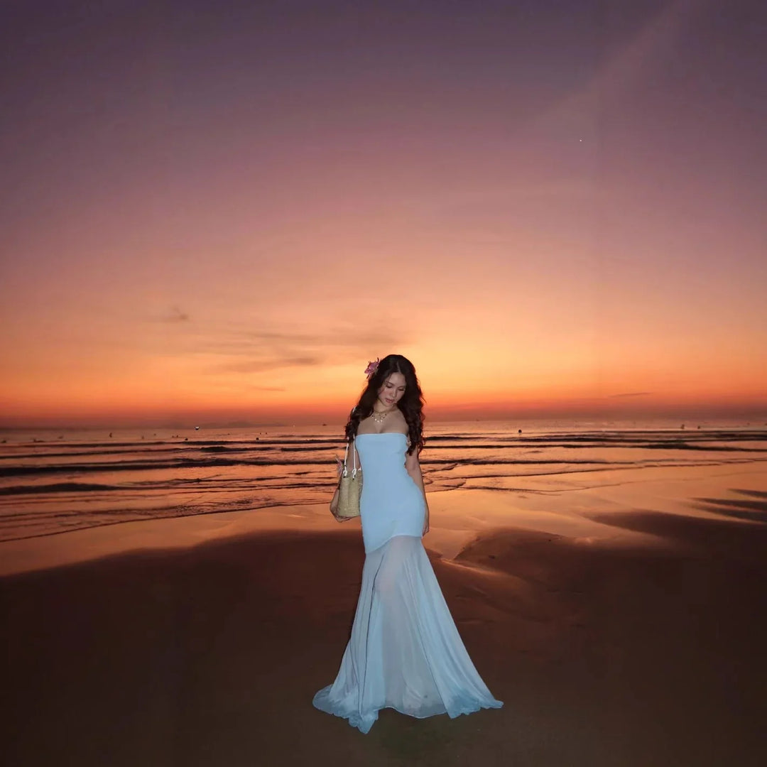 Woman in a flowing white dress standing on a beach at sunset with orange and purple sky
