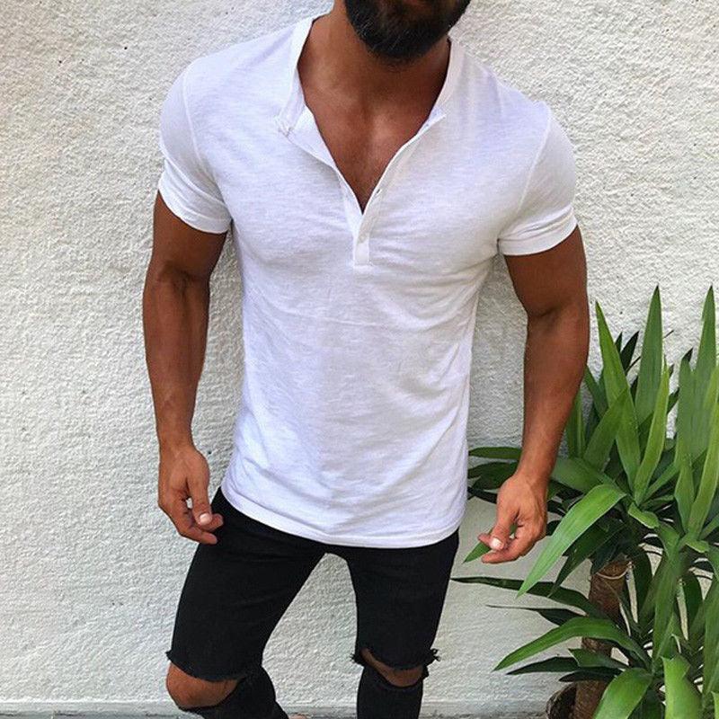 Muscular man wearing a fitted white henley shirt and ripped black jeans standing against white wall near green plant