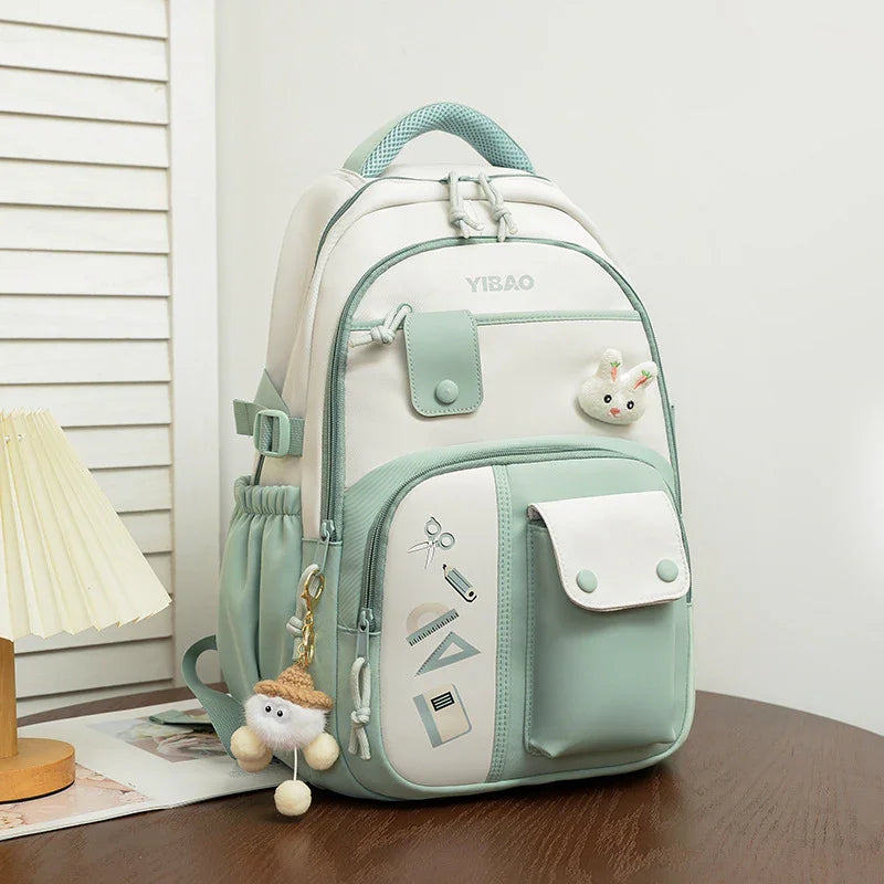 Light green and white Yibao backpack with front pockets, cute bunny pin, and fluffy keychain on wooden table