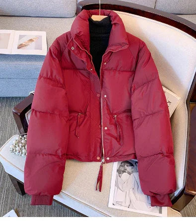 Red puffer jacket with zipper and snap buttons displayed on beige chair over black turtleneck sweater