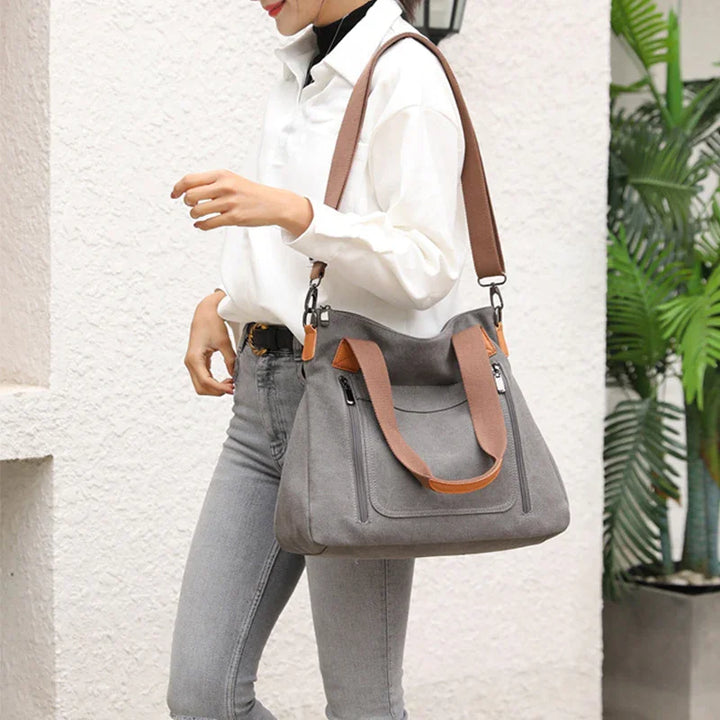 woman carrying gray canvas tote bag with brown straps, wearing white shirt and gray jeans, standing outdoors