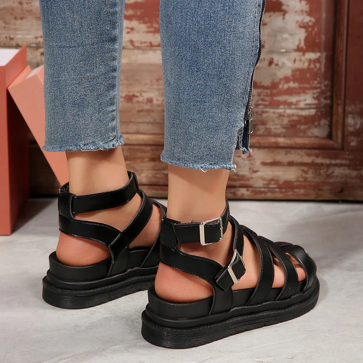 Back view of black strappy sandals with buckles on feet wearing frayed hem blue jeans against rustic wooden backdrop